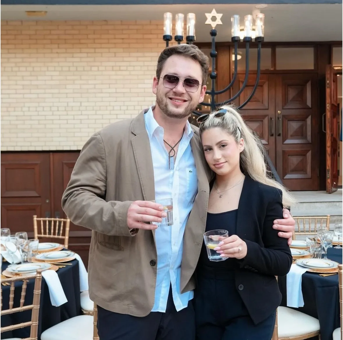 A young man and woman holding drinks pose outdoors by set dinner tables and a large menorah