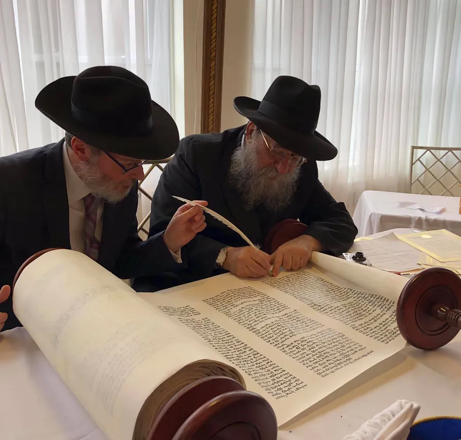 Two bearded men in black hats writing in a Torah scroll with a quill