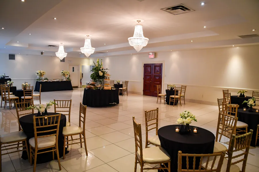 A white reception hall with crystal chandeliers, a tiled floor, wooden doors and round tables with gold chairs and white flowers