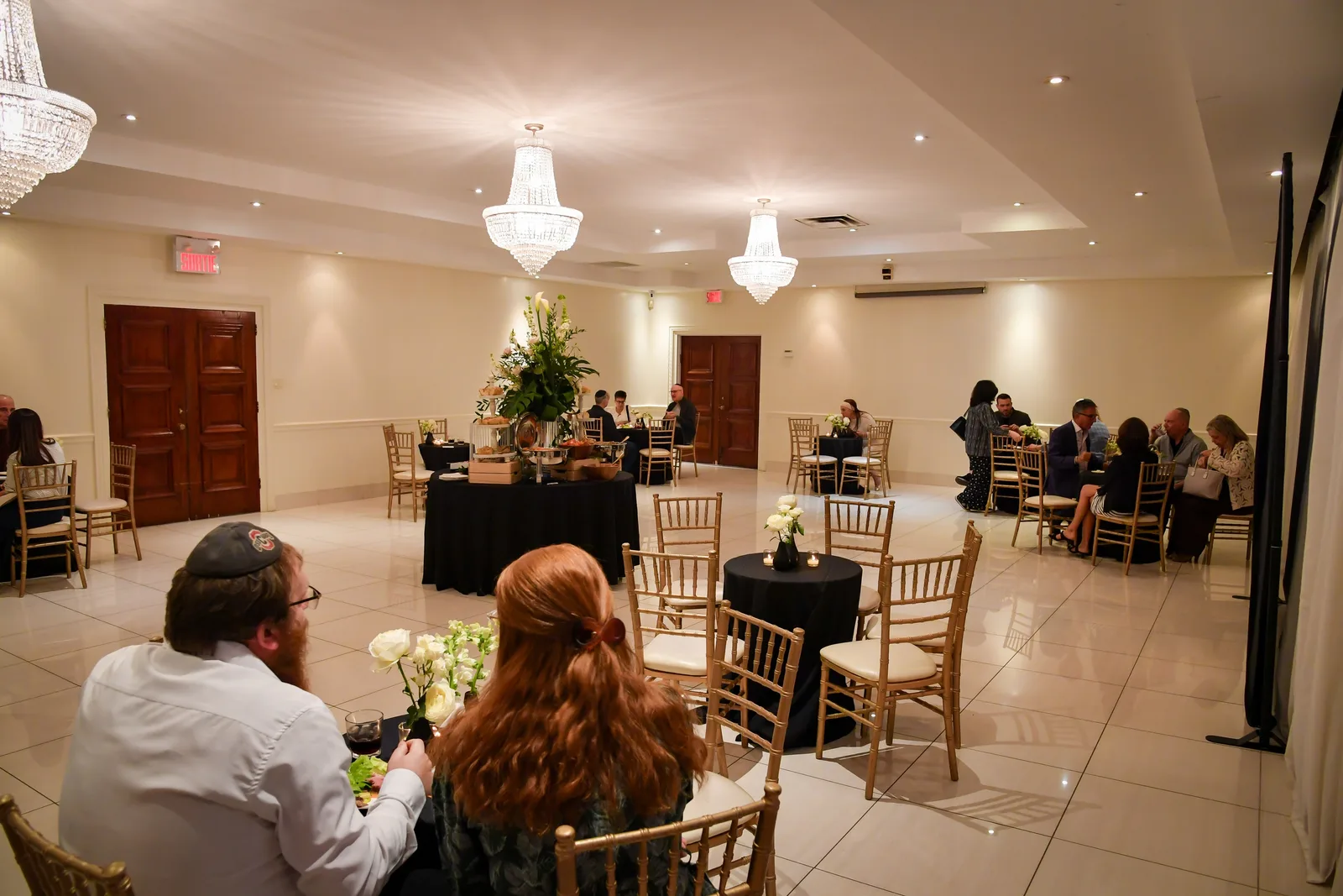 Guests chatting at small round tables in the white reception hall, under crystal chandeliers