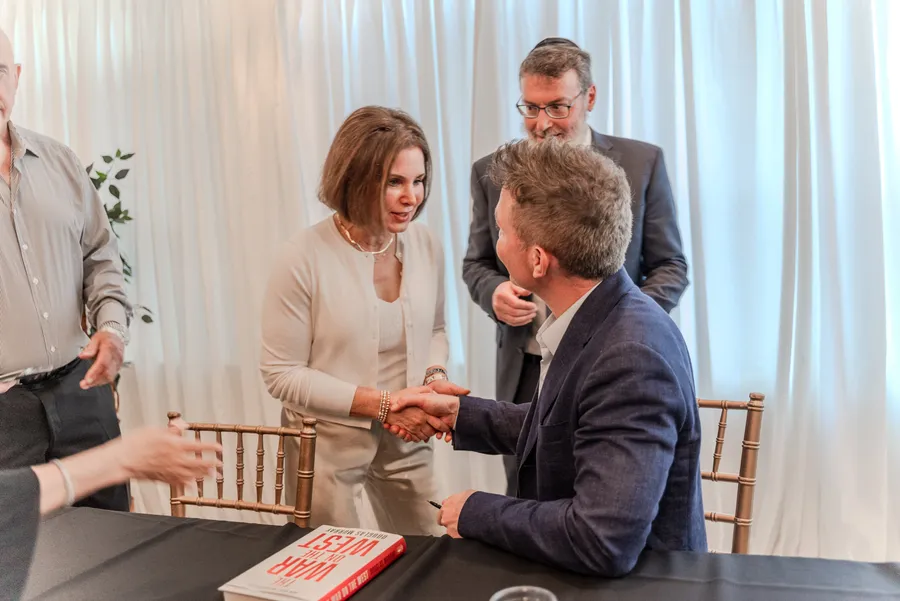 A woman shakes hands with a seated author at a signing table, a man in a kippah behind them