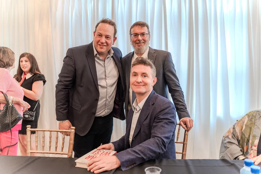 Two men in suits stand behind a smiling seated author with a book on the table