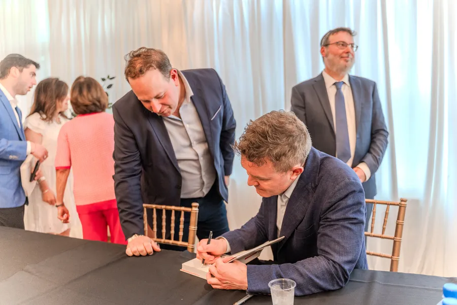 A man in a suit leans over the table as a seated author signs a book