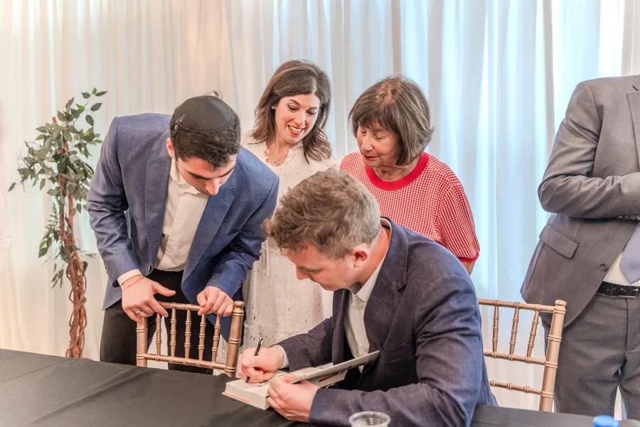 A seated author signs a book as a young man in a blue blazer and two women look on