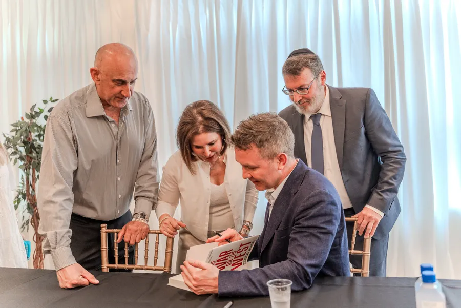 A seated author signs a book while a man and a woman lean in to watch
