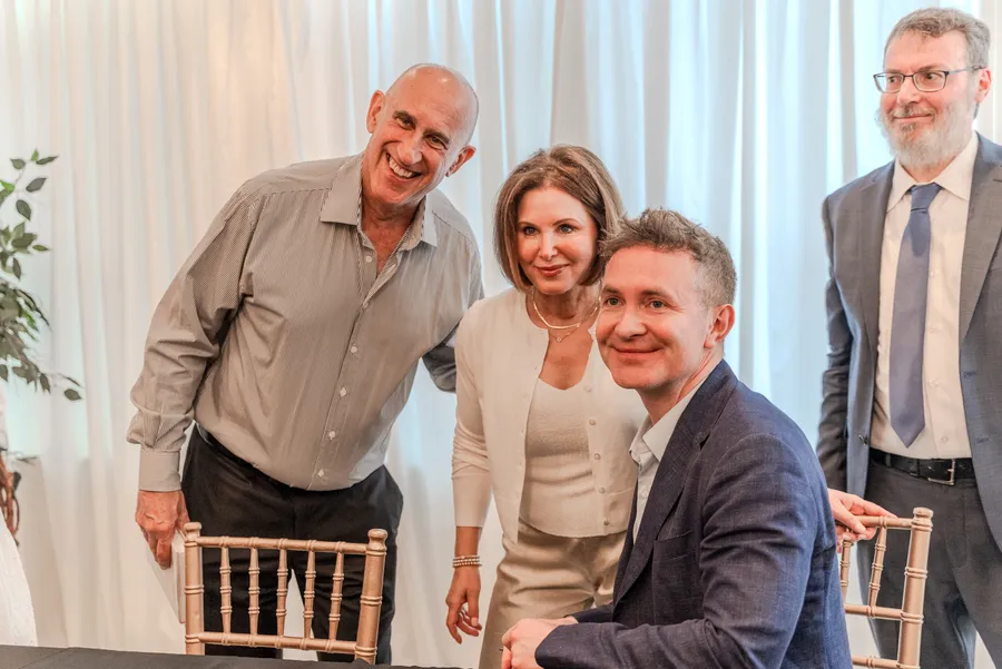 A smiling man and woman lean in beside a seated author at a signing table, a man in a suit nearby