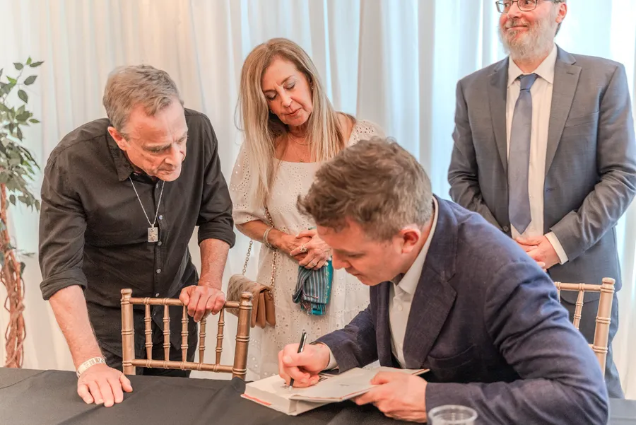 A seated author signs a book as a man and a woman lean on chairs to watch