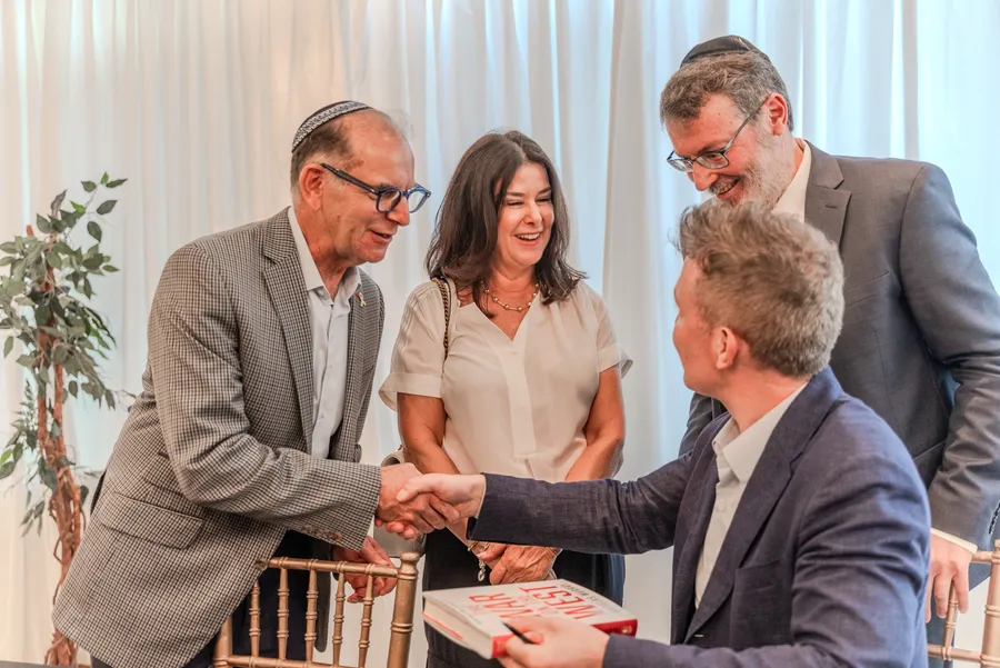 A man in a kippah shakes hands with a seated author across a table as others look on