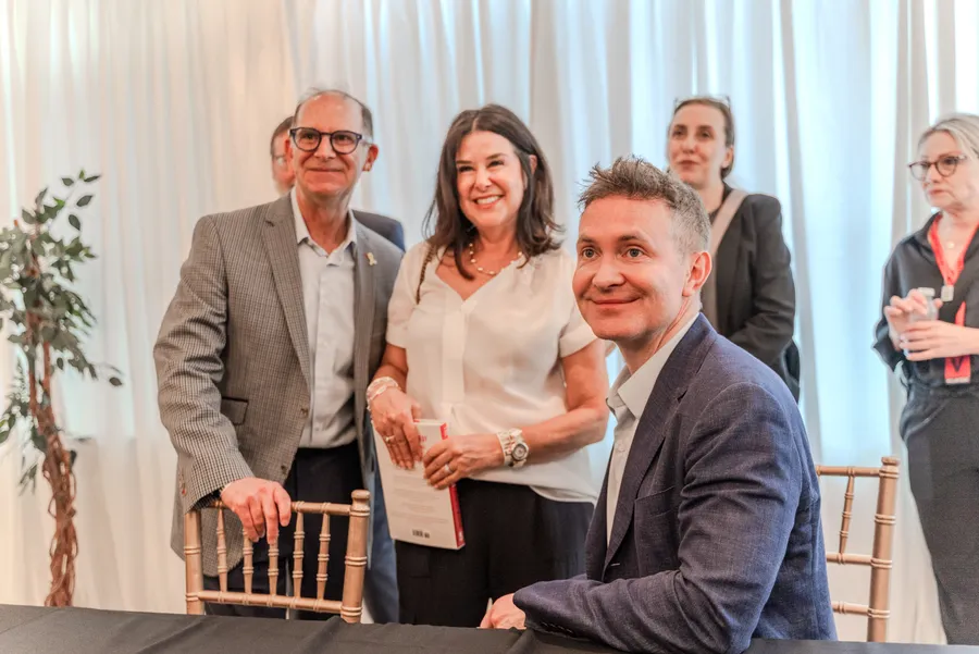 A man and a woman holding a book pose behind a smiling seated author at a signing table