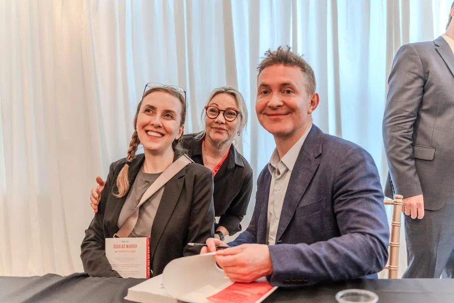 Two women pose beside a seated author at a signing table, one holding a red and white book
