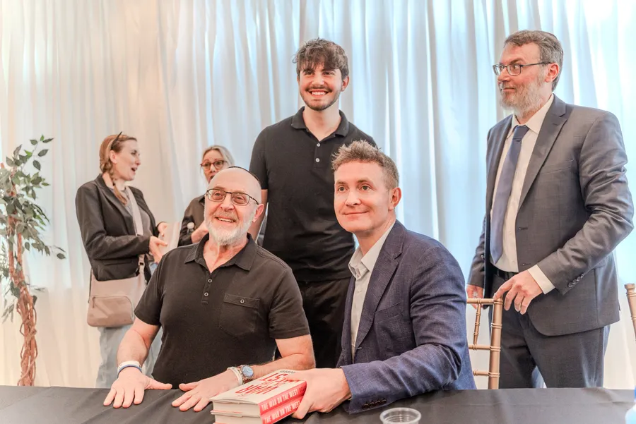 Five men smile together around a book signing table with a stack of red and white books