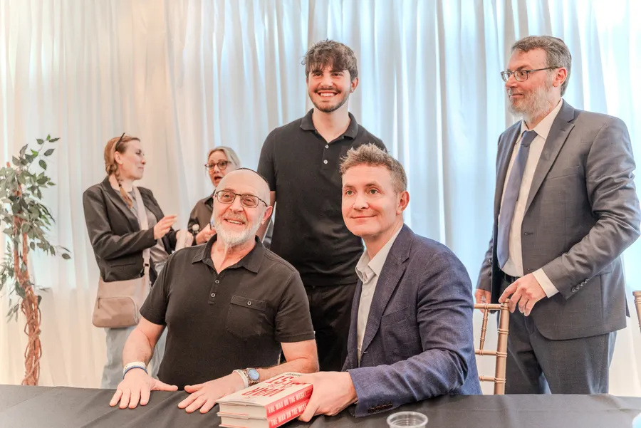 Five men pose around a signing table with a stack of red books, two women chatting in the background