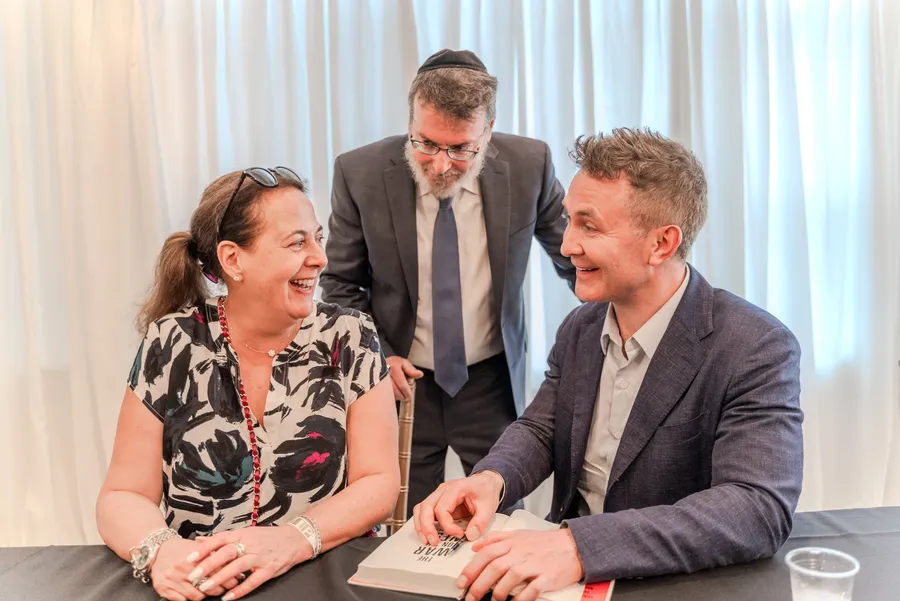 A woman in a patterned top laughs with a seated author at a signing table, a man in a kippah behind them