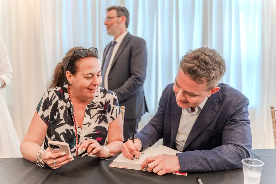A seated man signs a book as a woman in a patterned top holds her phone beside him