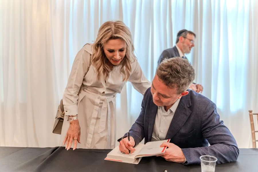 A seated man signs a book for a smiling woman in a beige coat while a man in a suit looks on