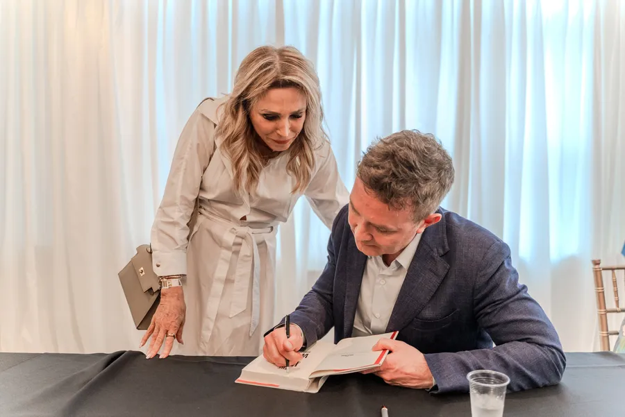 A man in a blue blazer signs a book at a table as a woman in a beige coat leans in to watch