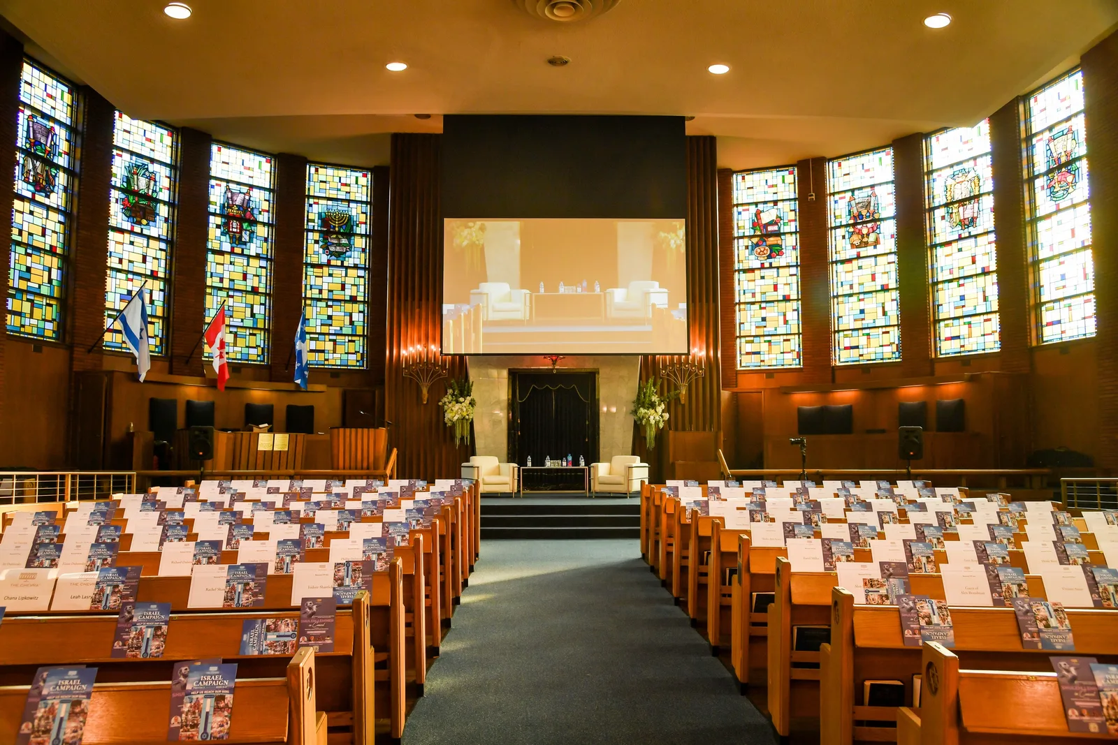 The empty sanctuary in daylight: the stained-glass windows lit up, wooden pews on both sides of the blue aisle, and the steps to the ark