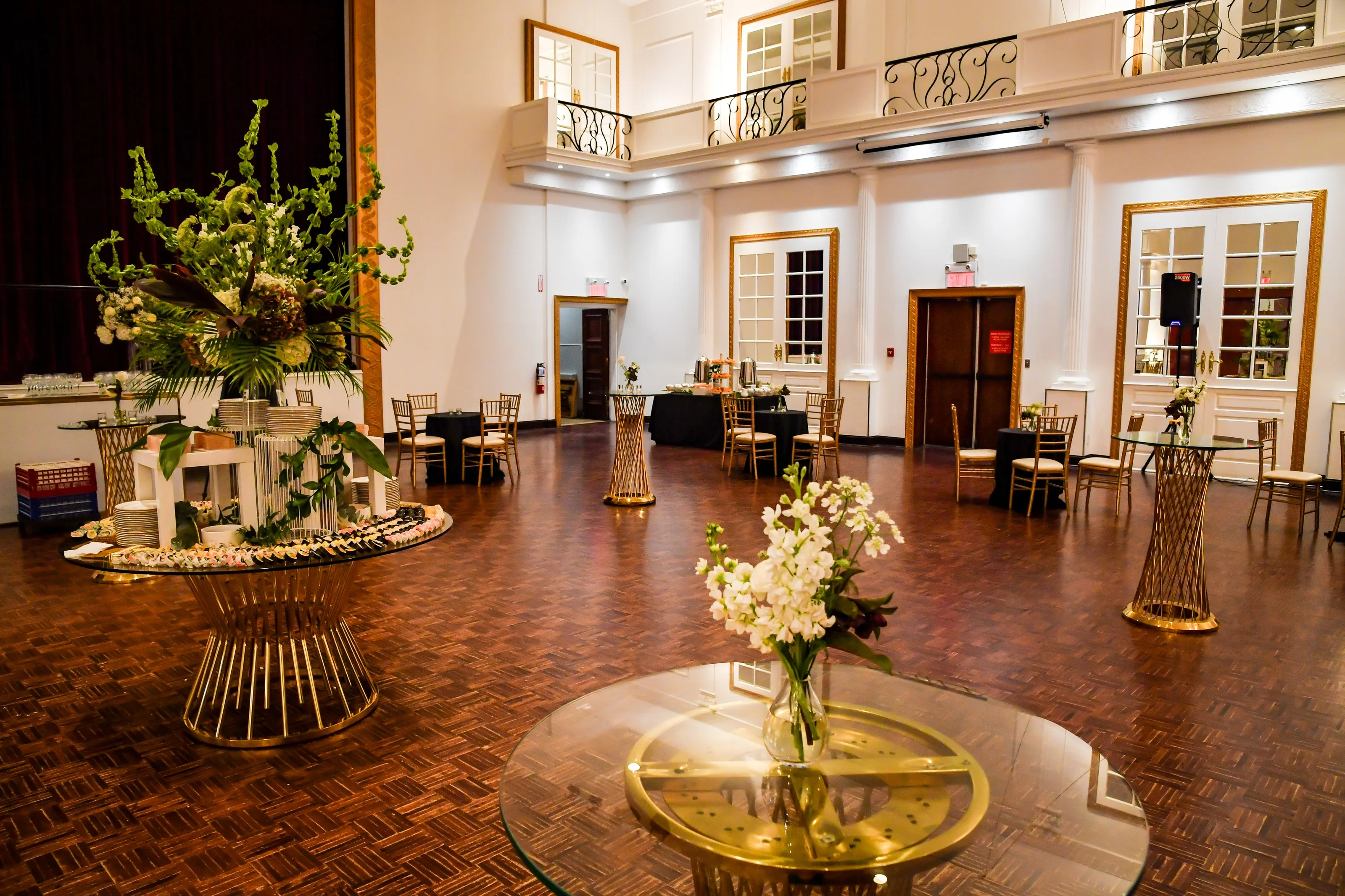 Singerman Hall set for a reception: Juliet balconies, French doors with gold frames, a parquet floor and tall flower arrangements