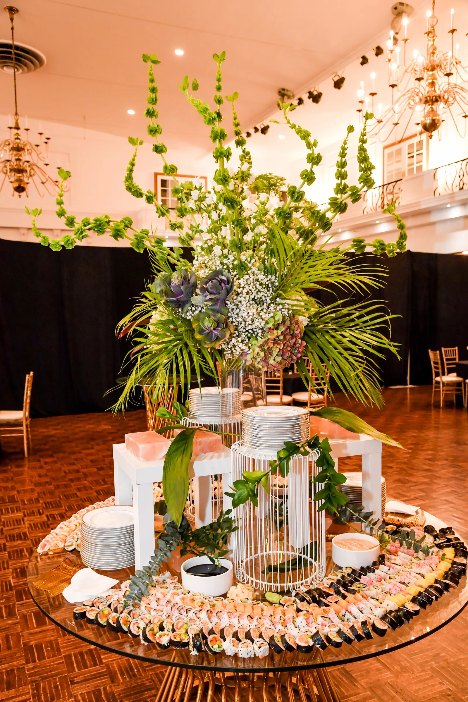 A round food station with a tall arrangement of greenery under the chandeliers in Singerman Hall