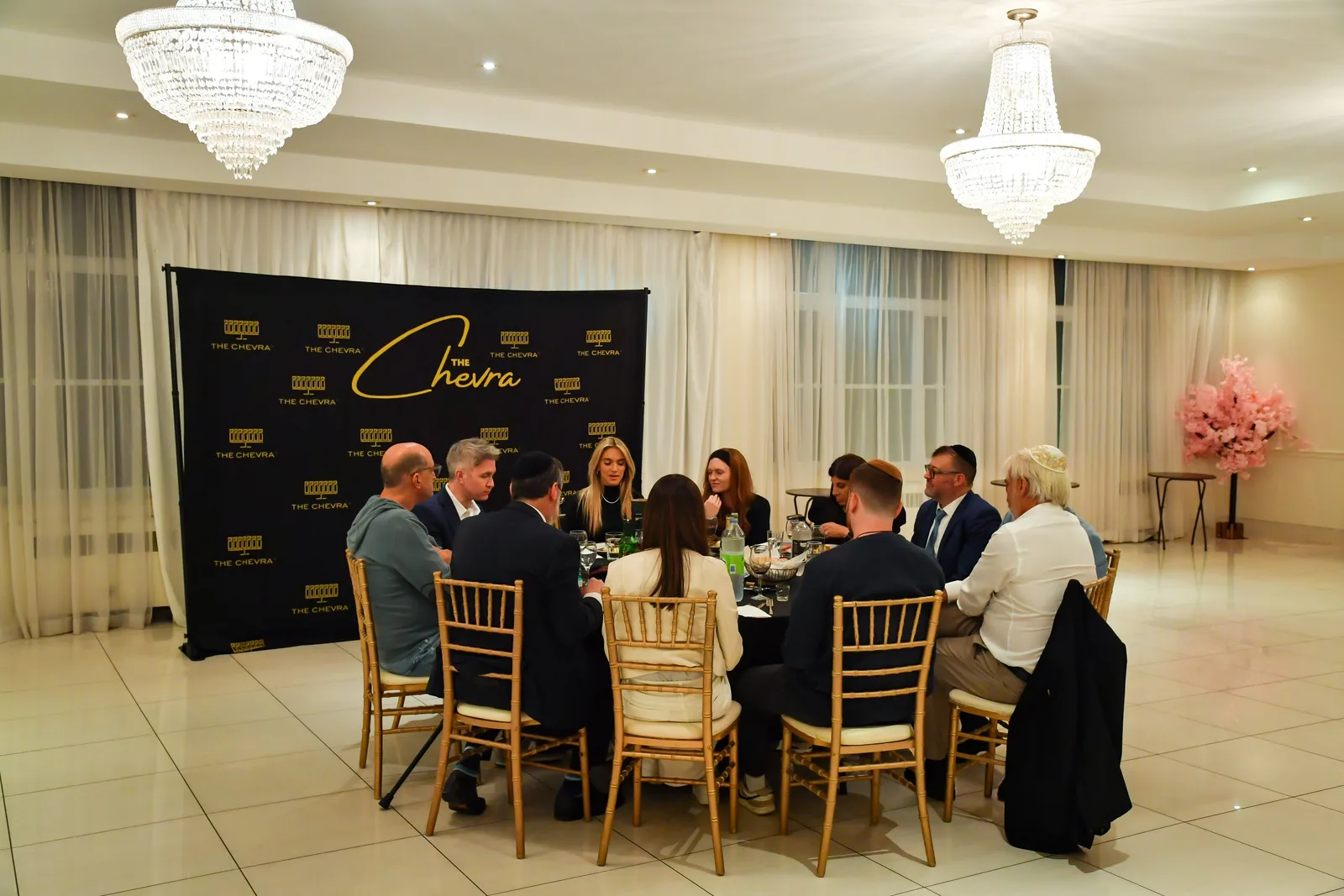 Guests around a dinner table in the white hall, gold chairs and crystal chandeliers
