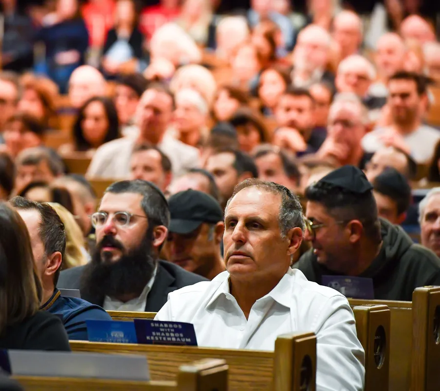A man in a white open-neck shirt listening in a full sanctuary beside a bearded man in round glasses; a card on the pew reads “Shabbos with Shabbos Kestenbaum”