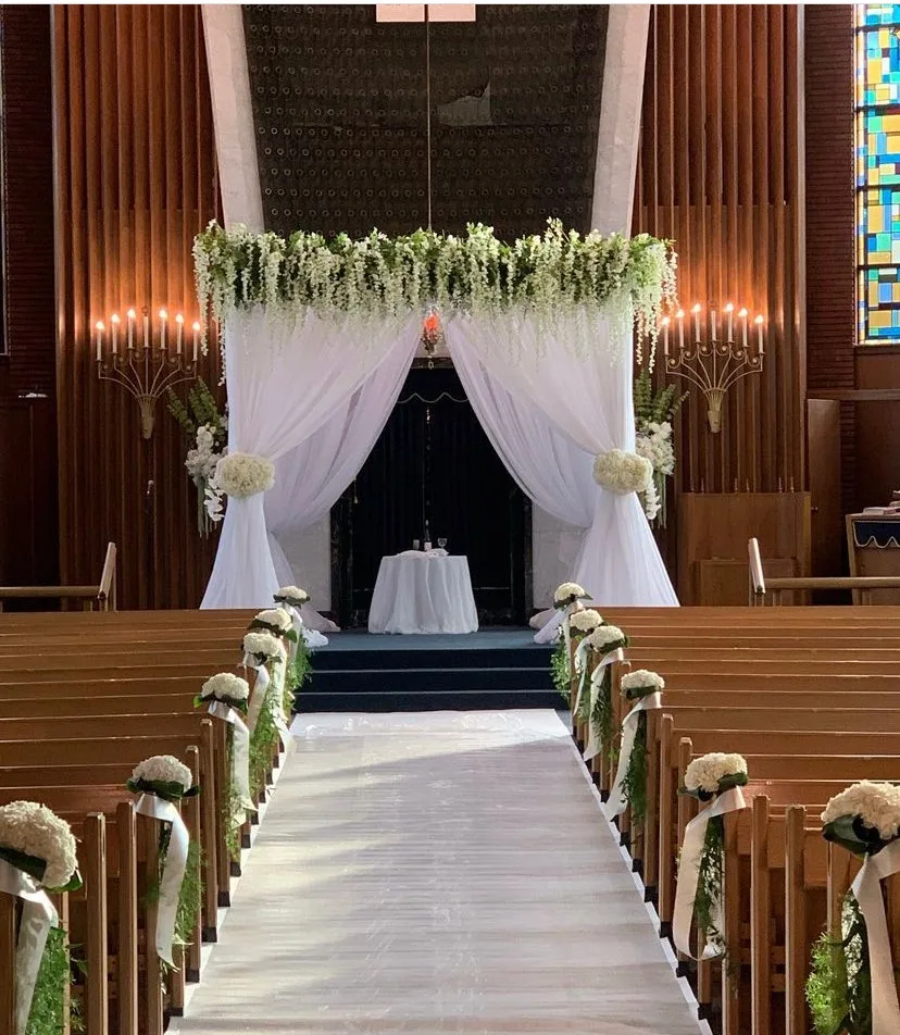 The sanctuary set for a wedding: a white aisle between the pews, white flowers on every pew end, and a chuppah of white drapes and hanging blossoms before the ark