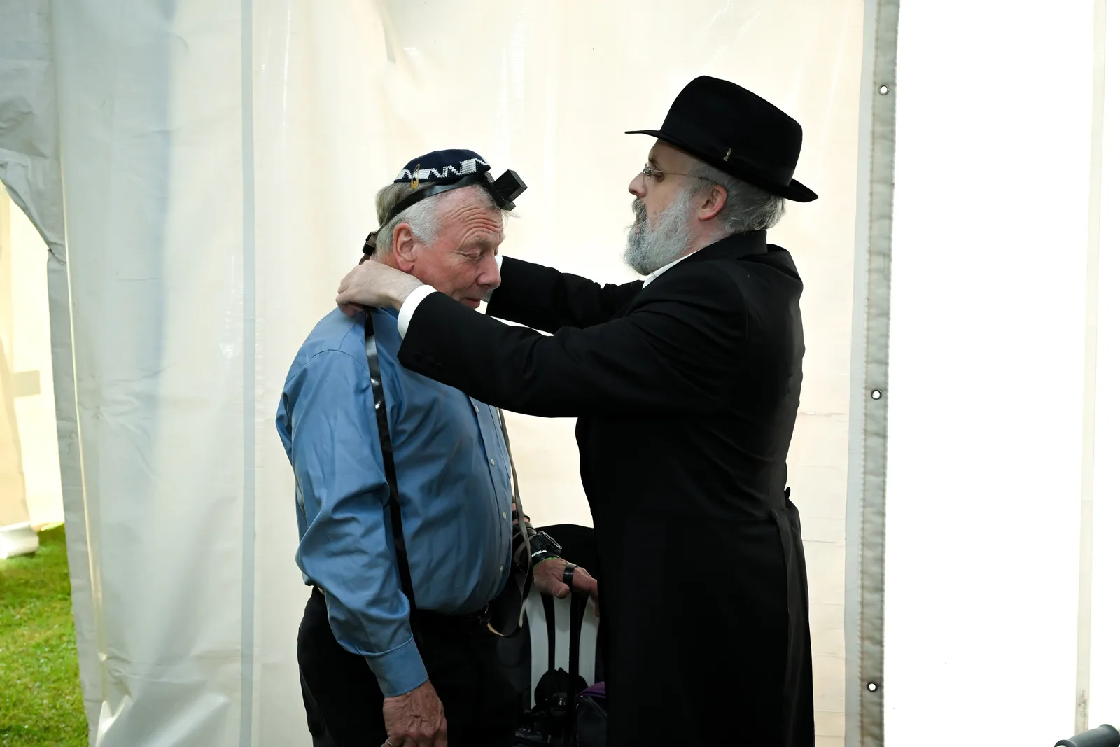 A rabbi in a black hat helps an older man put on tefillin, his hand resting on the man’s shoulder